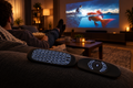 Black wireless air mouse remote with backlit keyboard on a sofa armrest with a movie projected on the wall in a cozy living room